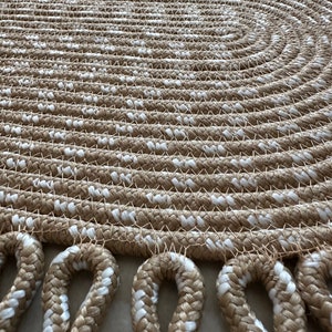 May include: A round, beige and white braided rug with a fringe border. The rug is made of thick rope and has a woven pattern.