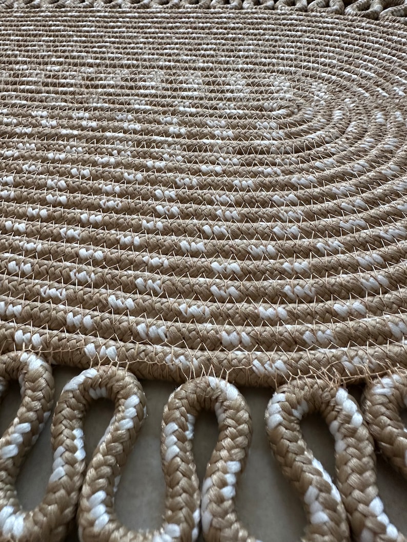 May include: A round, beige and white braided rug with a fringe border. The rug is made of thick rope and has a woven pattern.
