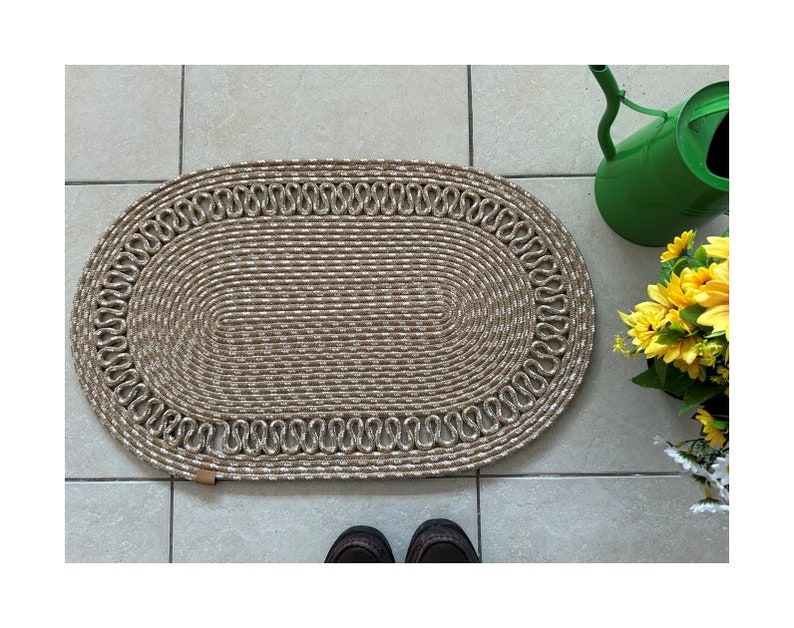 May include: A beige oval doormat with a braided rope design and a white trim. The doormat is on a tiled floor.