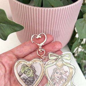 May include: A gold heart-shaped locket keychain with two separate compartments. The left compartment features a cartoon image of a woman with green skin and glasses. The right compartment features a cartoon image of a woman with blonde hair. The locket has a green ribbon bow.