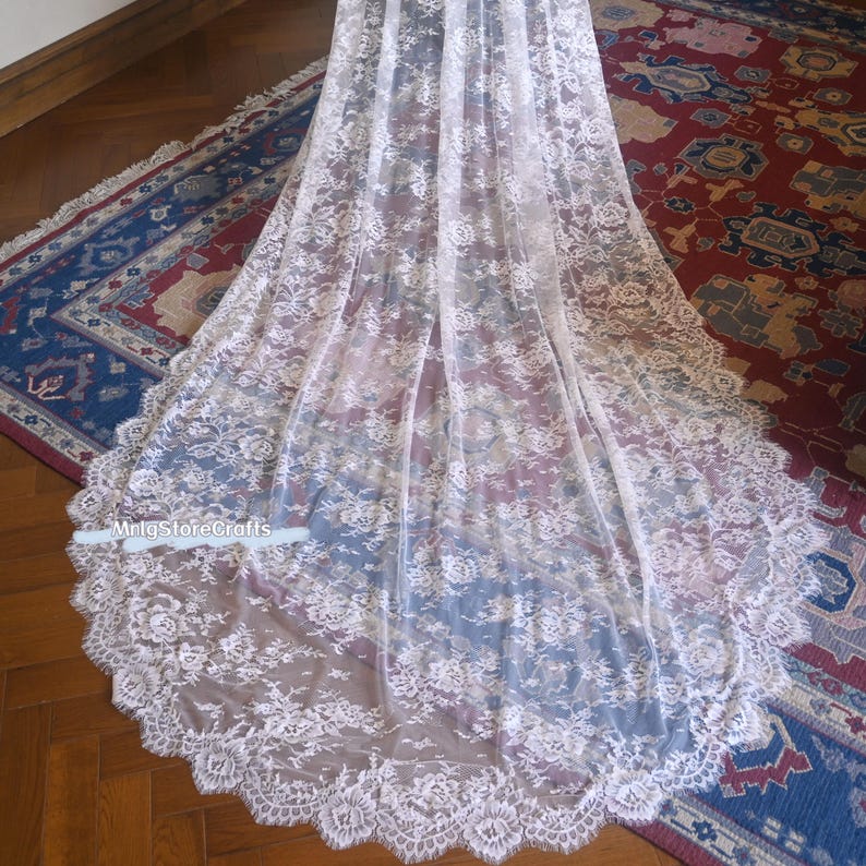May include: A long, white lace fabric with a floral pattern. The lace has a scalloped edge and is draped over a surface. The fabric is delicate and appears to be semi-transparent. The text "mrlgstorecrafts" is visible.