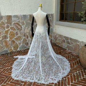 May include: A white lace wedding dress train displayed on a mannequin. The train features an intricate floral pattern and a scalloped edge. The mannequin has black arms and a gold-colored head. The dress is displayed on a brick patio.