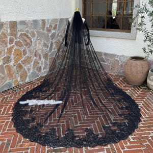May include: A long, black bridal veil with a detailed, floral lace trim. The veil is displayed on a mannequin, showcasing its length and intricate design. The veil is made of a sheer fabric.