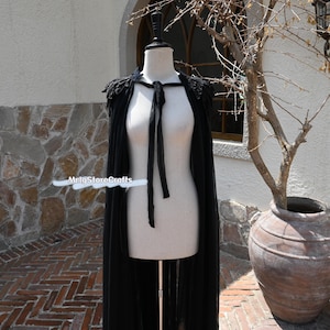 May include: A black sheer cape with lace detailing around the neckline and shoulders. The cape is tied at the neck with a black ribbon.