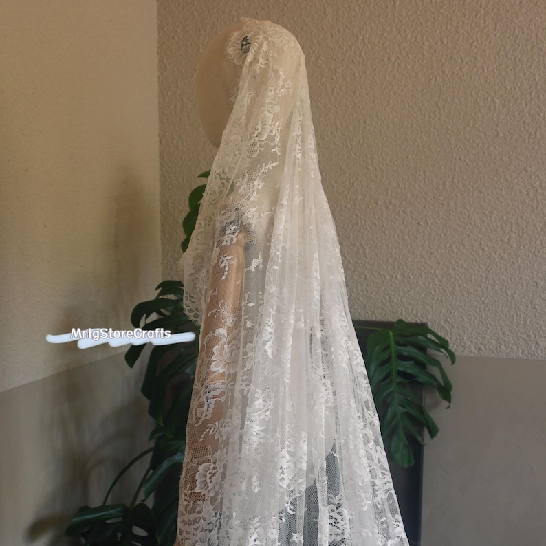 May include: A white lace veil with floral patterns, draped over a mannequin. The veil is long and flowing, with intricate details. The delicate lace creates a soft, romantic look, perfect for bridal wear.