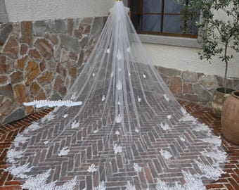 May include: A long, white bridal veil with intricate floral lace detailing. The veil features delicate, embroidered leaf and flower accents throughout, and a wide, ornate lace border along the hem. The veil is displayed on a mannequin, showcasing its full length and design.