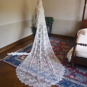 May include: A long, white lace wedding veil with intricate floral patterns. The veil is displayed on a mannequin, showcasing its length and delicate design. The veil's edge has a scalloped trim. The background includes a rug and a bed.