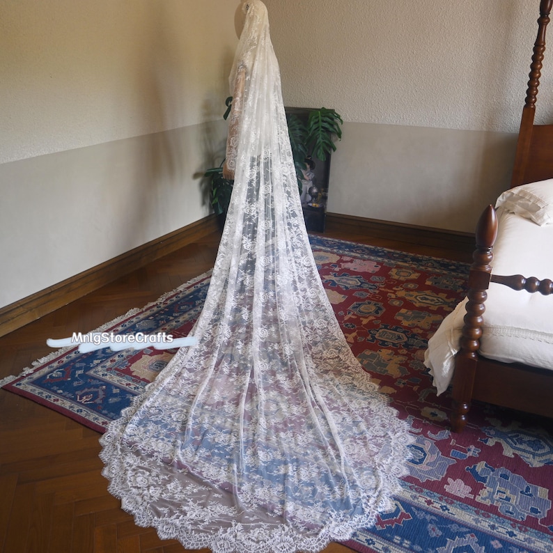 May include: A long, white lace wedding veil with intricate floral patterns. The veil is displayed on a mannequin, showcasing its length and delicate design. The veil's edge has a scalloped trim. The background includes a rug and a bed.