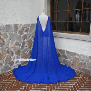 May include: A long, flowing royal blue cape with a deep V-back design. The cape is made of a lightweight, sheer fabric and drapes to the floor. The cape is displayed on a mannequin in front of a stone wall.