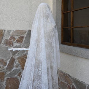 May include: A white lace veil, patterned with floral designs, draped over a mannequin. The sheer fabric is displayed near a window and a stone wall. The text "MrigStoreCrafts" is visible. The overall effect is soft and romantic.
