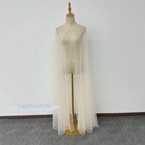 May include: A sheer ivory tulle cape with a simple design, displayed on a mannequin. The cape is long and flowing, with a slight shimmer.