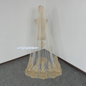 May include: A long ivory bridal veil with a gold lace trim. The lace features a floral design with intricate details.