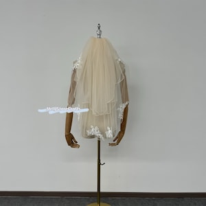 May include: A single-tier ivory bridal veil with delicate white lace trim. The veil is displayed on a wooden mannequin.