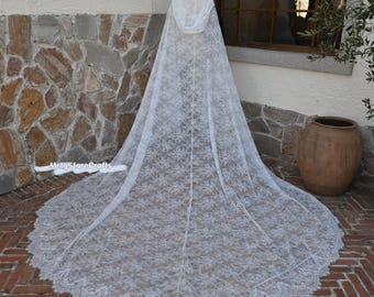 May include: A white lace hooded cape with a long train. The cape is made of delicate floral lace and has a scalloped edge. The cape is displayed on a mannequin in front of a stone wall.