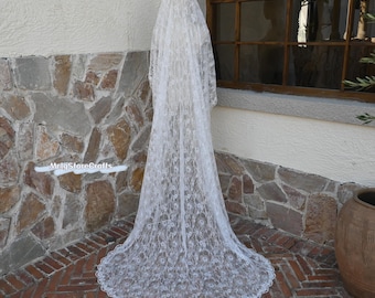 May include: A long, white lace wedding veil with a floral pattern. The veil is displayed outdoors, draped against a stone wall and brick floor. The veil's delicate lace design is visible, with a scalloped edge.