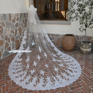 Op de afbeelding: Een lange, witte bruidssluier met ingewikkelde bloemenkantdetails langs de randen en verspreid over de transparante stof. De sluier wordt op een mannequin getoond, waarbij de lengte en het ontwerp worden benadrukt. De achtergrond is een bakstenen terras en een stenen muur.