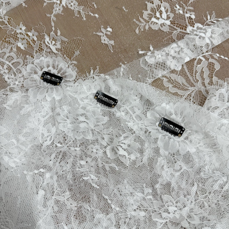 May include: Close-up of white lace fabric with floral patterns. Three black hair clips are attached to the lace. The lace is delicate and detailed, with a variety of floral designs.