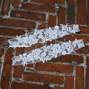 May include: Two white floral lace appliques with intricate detailing. The appliques feature a delicate floral pattern with sequins, perfect for embellishing bridal wear or other craft projects. The appliques are displayed against a brick wall.