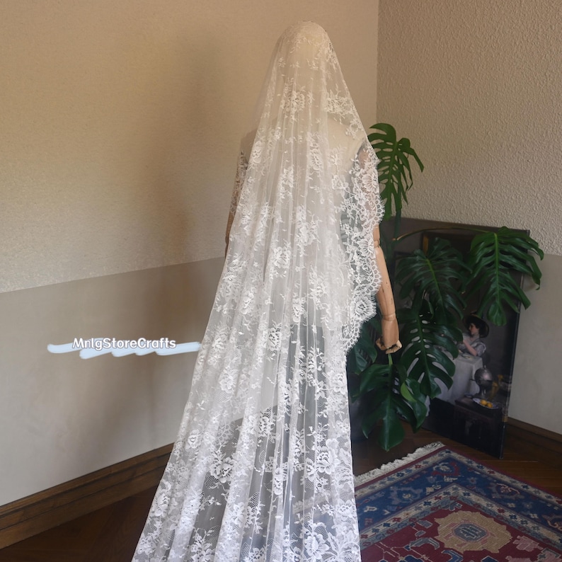 May include: A long, white lace bridal veil with intricate floral patterns. The veil is draped over a mannequin, showcasing its delicate design and flowing fabric. The veil's edges are finished with a scalloped lace trim.