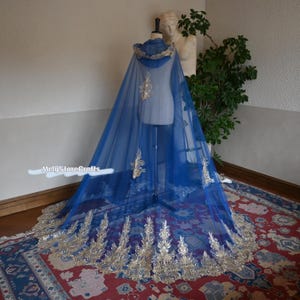 May include: A royal blue cape with a hood and gold embroidery. The cape is displayed on a mannequin, showcasing its flowing design and intricate details. The bottom edge features elaborate gold leaf patterns.
