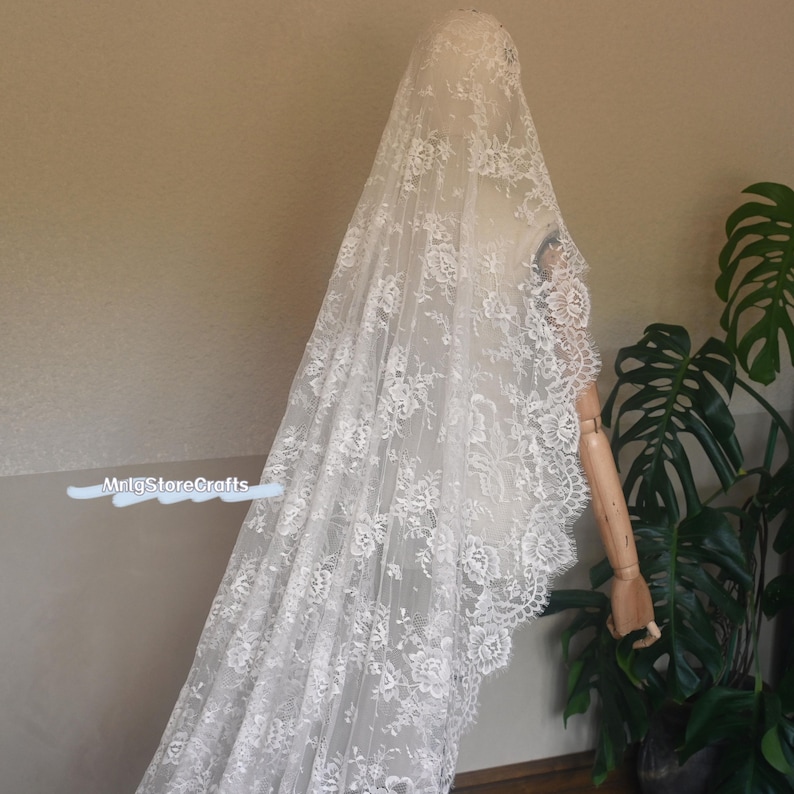 May include: A long, white lace veil with floral patterns, draped over a mannequin. The veil's edge is scalloped, and the delicate lace creates a soft, romantic look. The veil is displayed against a neutral background with a plant.