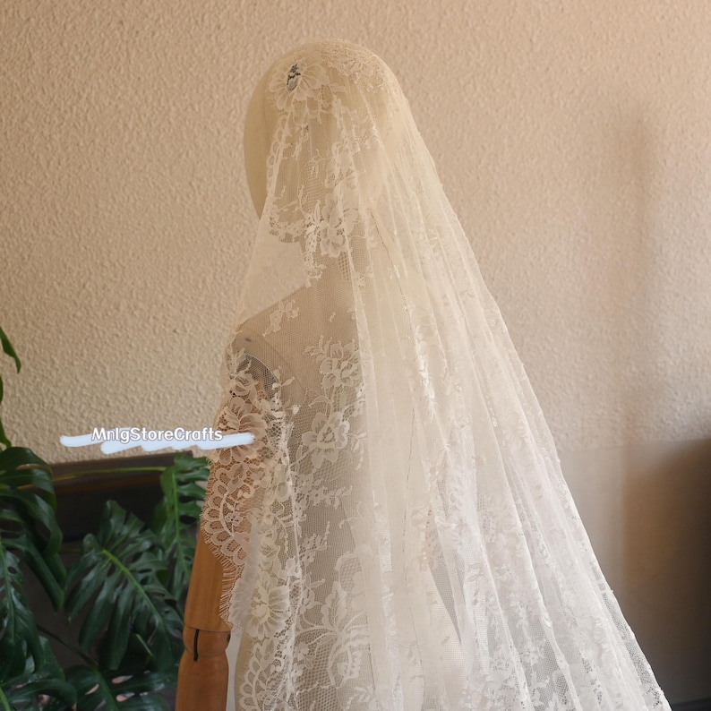 May include: Ivory lace bridal veil draped over a mannequin. The veil features intricate floral patterns along the edges and a delicate, sheer fabric. The veil is long and flowing, creating a romantic and elegant look. The MrIgStoreCrafts watermark is visible.