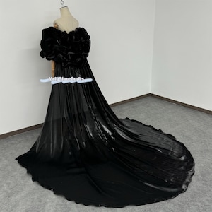 Puede incluir: Un vestido de maternidad negro con un corpiño grande y con volantes y una falda larga y fluida. El vestido está hecho de una tela suave y transparente con un acabado satinado.