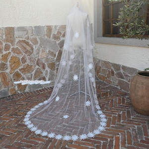 Op de afbeelding: Een lange, witte bruidssluier met bloemenkanten applicaties en verspreide parelaccenten. De sluier wordt getoond op een mannequin, die het delicate ontwerp en de ingewikkelde details laat zien. De rand van de sluier is versierd met een herhalend bloemenpatroon.