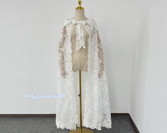 May include: White lace bridal cape with a delicate floral pattern. The cape is draped over a mannequin and has a bow at the neck.