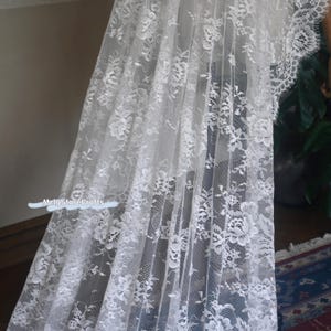 May include: A white lace fabric with a floral pattern. The delicate lace is draped, showcasing the intricate design and texture. The fabric is semi-transparent, allowing light to filter through, creating a soft, airy appearance. The overall aesthetic is elegant and romantic.