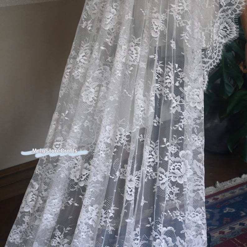 May include: A white lace fabric with a floral pattern. The delicate lace is draped, showcasing the intricate design and texture. The fabric is semi-transparent, allowing light to filter through, creating a soft, airy appearance. The overall aesthetic is elegant and romantic.
