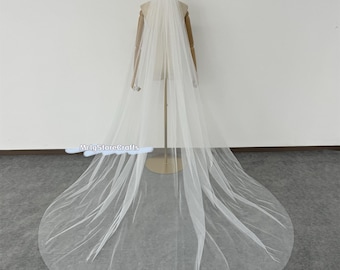 May include: A long, white tulle wedding veil with a single layer of soft, sheer fabric. The veil is draped over a mannequin and falls to the floor in a graceful cascade.