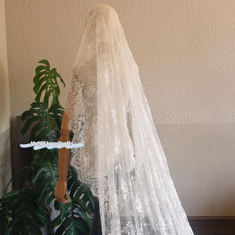 May include: Ivory-colored lace veil, draped over a mannequin. The veil features intricate floral patterns and a long, flowing design. The delicate lace creates a soft, romantic look, perfect for bridal wear. The veil is displayed against a neutral background.