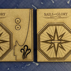 Puede incluir: Dos piezas de cart&oacute;n marr&oacute;n para un juego, una con una rosa de los vientos y la otra con una rosa de los vientos y un c&iacute;rculo con un dise&ntilde;o de estrella. El texto "SAILS of GLORY NAPOLEONIC WARS" est&aacute; impreso en ambas piezas.