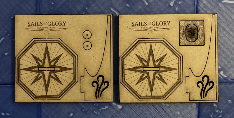 Puede incluir: Dos piezas de cart&oacute;n marr&oacute;n para un juego, una con una rosa de los vientos y la otra con una rosa de los vientos y un c&iacute;rculo con un dise&ntilde;o de estrella. El texto "SAILS of GLORY NAPOLEONIC WARS" est&aacute; impreso en ambas piezas.