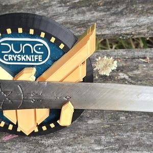 May include: A silver knife with a brown wooden handle and a decorative design. The knife is mounted on a black and gold display stand with the text "Dung CrySknife".
