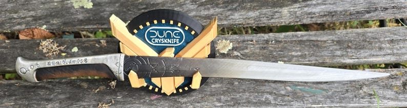 May include: A silver knife with a brown wooden handle and a decorative design. The knife is mounted on a black and gold display stand with the text "Dung CrySknife".