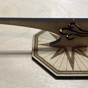 Puede incluir: Una br&uacute;jula de madera con un dise&ntilde;o decorativo y una aguja m&oacute;vil. La br&uacute;jula est&aacute; hecha de madera marr&oacute;n claro y tiene un borde negro. La aguja est&aacute; hecha de madera marr&oacute;n oscuro y tiene una punta negra.