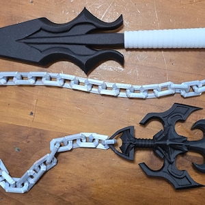 Pode incluir: Duas armas de lâmina pretas e brancas conectadas por uma corrente prateada. A arma superior tem uma empunhadura branca e uma lâmina preta com bordas decorativas. A arma inferior é toda preta com um design semelhante.
