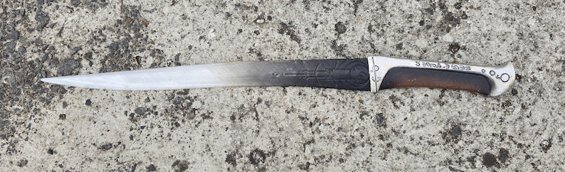 May include: A silver and black dagger with a wooden handle. The blade has a decorative pattern and the handle has the text "S. MATTHIAS. A.D. 1538".