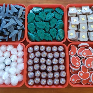 Może przedstawiać: Sześć czerwonych plastikowych pojemników wypełnionych różnymi kolorowymi elementami gry. Pojemniki są ułożone w dwóch rzędach po trzy. Elementy obejmują szare metalowe elementy, zielone liście, beżowe kawałki chleba, białe elementy w kształcie diamentu, brązowe okrągłe elementy i czerwone elementy mięsne.
