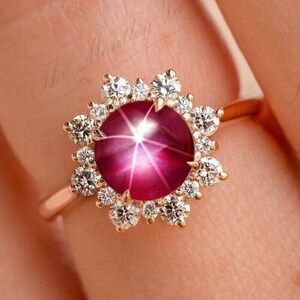 May include: A rose gold ring featuring a large, round, star ruby gemstone surrounded by a halo of small, clear diamonds. The ruby has a deep pink hue, and the diamonds sparkle against the rose gold band. The ring is set on a finger.