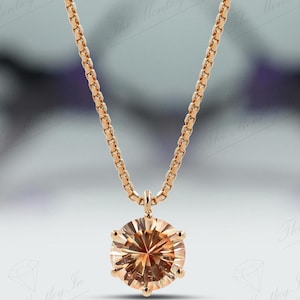 May include: A gold-colored necklace with a round, light brown gemstone pendant. The pendant is faceted, catching the light. The chain is a delicate box chain style. The background is blurred, with hints of purple and white.