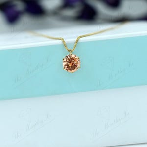 May include: A gold necklace with a peach-colored gemstone pendant. The pendant is oval-shaped and set in a gold setting. The necklace is displayed on a white and light blue box.