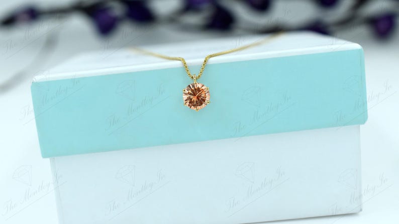 May include: A gold necklace with a peach-colored gemstone pendant. The pendant is oval-shaped and set in a gold setting. The necklace is displayed on a white and light blue box.