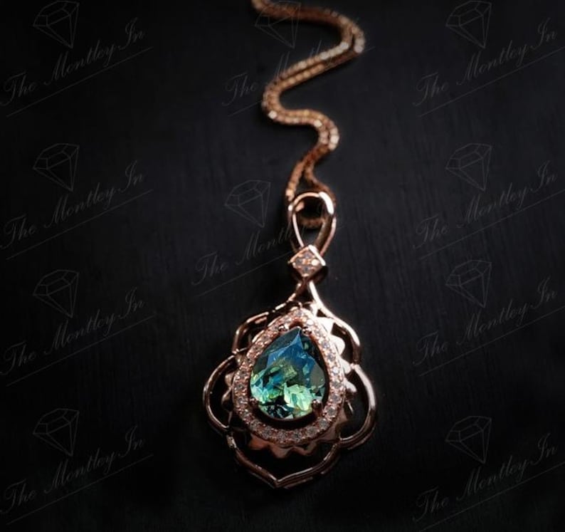 Puede incluir: Un collar con colgante de oro rosa con una piedra preciosa azul-verdosa en forma de l&aacute;grima. El colgante est&aacute; adornado con peque&ntilde;as piedras transparentes y filigrana de oro rosa. La cadena tambi&eacute;n es de oro rosa, sobre un fondo negro.