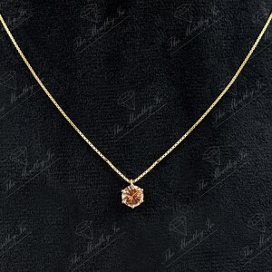 May include: A delicate gold necklace with a small, round, light brown gemstone pendant. The pendant is set in a gold bezel and hangs from a thin gold chain. The background is a dark, textured surface.