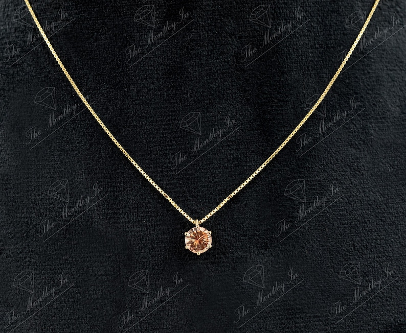 May include: A delicate gold necklace with a small, round, light brown gemstone pendant. The pendant is set in a gold bezel and hangs from a thin gold chain. The background is a dark, textured surface.