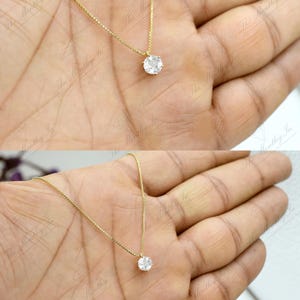 Pu&ograve; includere: Una delicata collana d'oro con un singolo pendente rotondo e trasparente con pietra preziosa. Il pendente &egrave; sospeso a una sottile catena d'oro. La collana &egrave; mostrata su una mano.