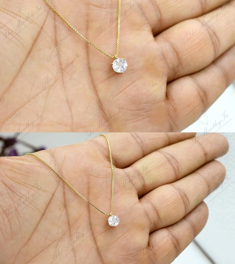 Pu&ograve; includere: Una delicata collana d'oro con un singolo pendente rotondo e trasparente con pietra preziosa. Il pendente &egrave; sospeso a una sottile catena d'oro. La collana &egrave; mostrata su una mano.